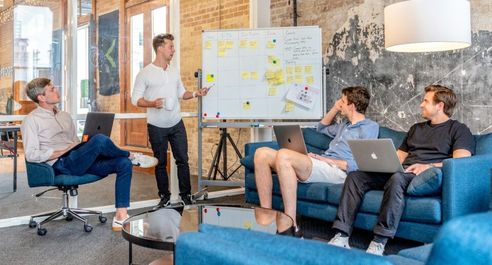 three men sitting while using laptops and watching man beside whiteboard